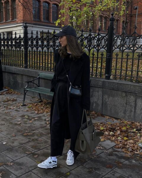 Kobieta w modnej, miejskiej stylizacji spaceruje po chodniku wzdłuż budynku. Ma na sobie czarne legginsy, biały T-shirt, szarą, oversize'ową marynarkę oraz białe skarpetki. Na głowie ma czarną czapkę z daszkiem z białym logo (NY) i okulary przeciwsłoneczne. W ręku trzyma czarną torebkę typu półksiężyc. Na nogach ma srebrno-białe sneakersy New Balance w stylu retro.