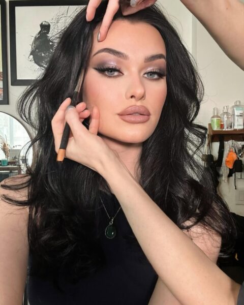 Zdjęcie przedstawia wizażystkę, która precyzyjnie poprawia brwi modelki, koncentrując się na perfekcyjnym wykończeniu cery oraz intensywnym makijażu oka. Smoky eye zostało wykonane w chłodnych odcieniach fioletu i szarości z błyszczącym, metalicznym akcentem na powiece. Taki dopracowany, glamour look z mocno zarysowanymi ustami nude idealnie wpasowuje się w bardziej wieczorowe trendy w makijażu 2026.