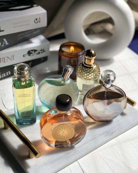 Kilka flakonów perfum o różnych kształtach i kolorach ustawionych na jasnej tacy obok zapalonej świecy i książek. Kompozycja tworzy estetyczną, domową scenę, w której zapachy są wyeksponowane w świetle dziennym. To przykład aranżacji, która wygląda efektownie, ale wymaga ostrożności przy dłuższym przechowywaniu perfum.