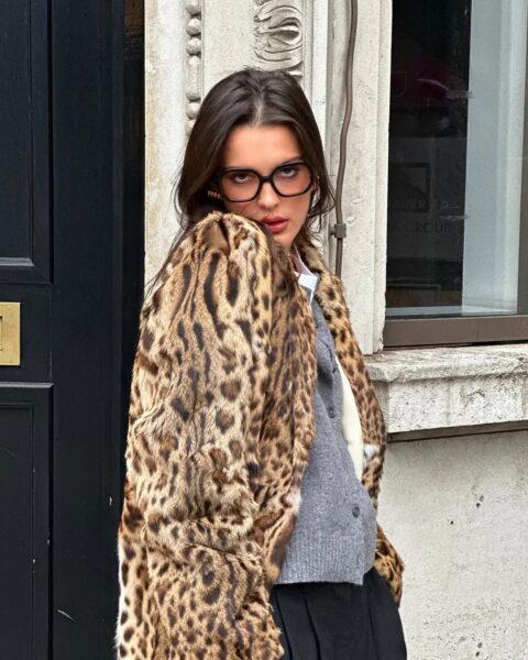 Zbliżenie na twarz i tors modelki w futrzanym płaszczu o wyrazistym, cętkowanym wzorze leoparda, który będzie prawdopodobnie jednym z trendów na zimę powracających do mody zwierzęcej. Pod futrem widoczny jest szary, wełniany kardigan noszony na białej koszuli oraz czarna, plisowana spódnica, co nadaje całości szykownego charakteru, a duże okulary w bursztynowej oprawie dopełniają ten vintage'owy i zarazem modny zestaw.