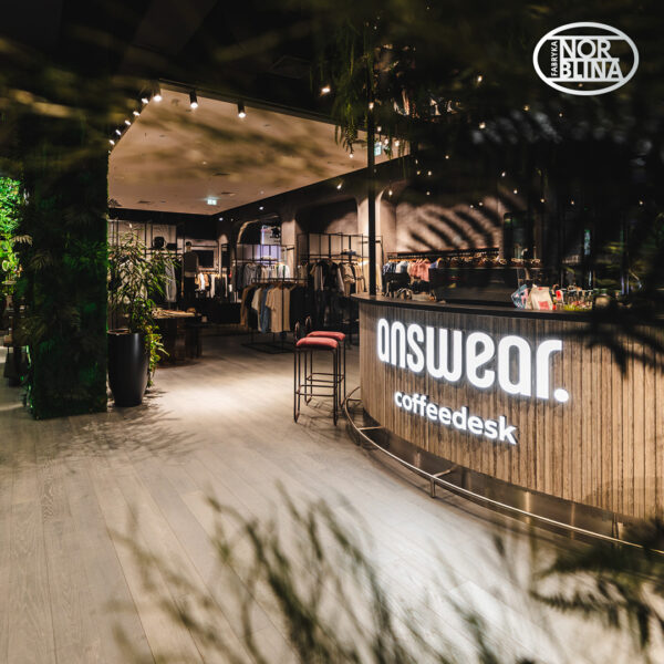 Wnętrze Answear Concept Store w Fabryce Norblina z widocznym neonem przy ladzie i selekcją ubrań na wieszakach. To jedno z warszawskich concept store'ów do szukania prezentów, które łączą modę z przyjazną, nowoczesną przestrzenią. Rośliny i oświetlenie tworzą tu lekki, butikowy klimat.
