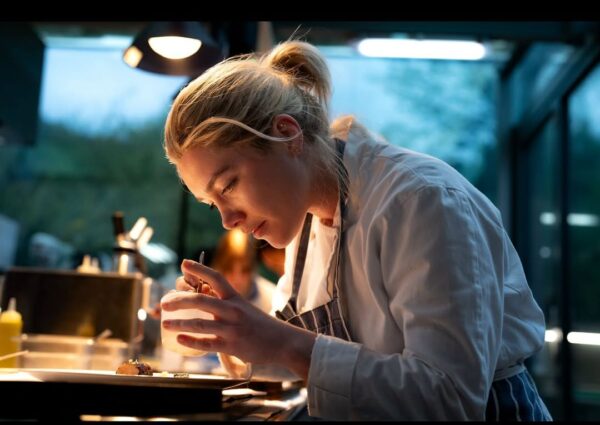 Zbliżenie na kobietę o jasnych włosach, związaną w kucyk, ubraną w biały kitel i pasiasty fartuch, która z pełnym skupieniem dekoruje danie w nowoczesnej, nieco mrocznej kuchni. Intensywna praca w gastronomii i ten pełen detali kadr przypominają mi o dramatyzmie, który często towarzyszy produkcjom pretendującym do miana najlepszego filmu 2025 — zwłaszcza tym opowiadającym o pasji i wyzwaniach.
