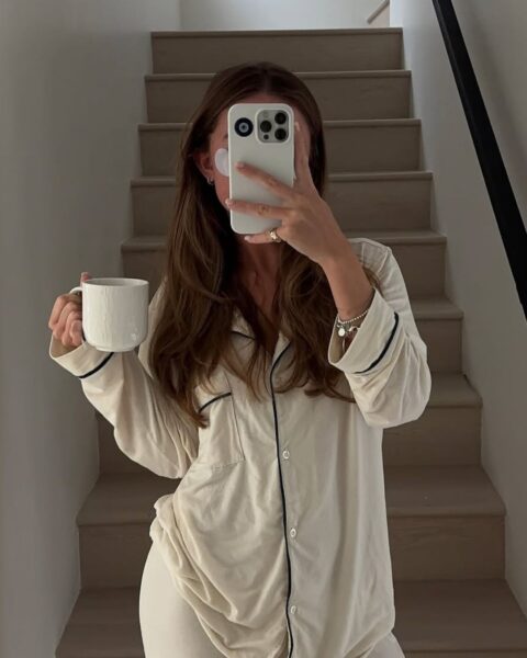 Selfie zrobione na klatce schodowej, ukazujące kobietę w kremowej piżamie z ciemną lamówką. Trzyma ona biały kubek, a jej twarz jest częściowo zasłonięta telefonem. Na policzku widoczna jest maseczka pod oczy, co podkreśla rutynę pielęgnacyjną. Ten klasyczny, a jednocześnie wygodny fason demonstruje, jak luksusowe piżamy na 2026 rok z naturalnych materiałów łączą poranną pielęgnację z domowym szykiem.