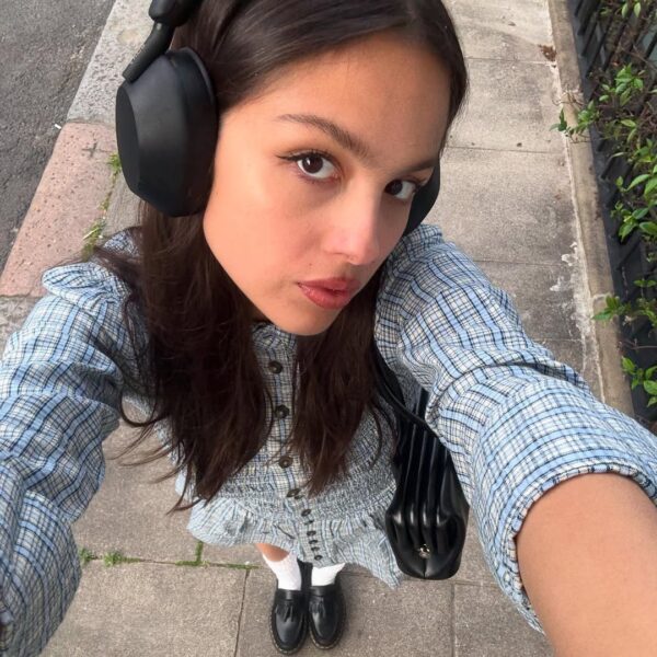 Selfie wykonane z góry przedstawia młodą kobietę na chodniku, która uosabia styl trendy Gen Z – ma na sobie duże, czarne słuchawki, kratkowaną, niebiesko-szarą sukienkę w stylu cottagecore i czarne mokasyny na białych skarpetkach. Trzyma dużą, czarną torebkę na ramieniu, a jej intensywne spojrzenie skierowane jest prosto w obiektyw, oddając współczesny uliczny klimat.