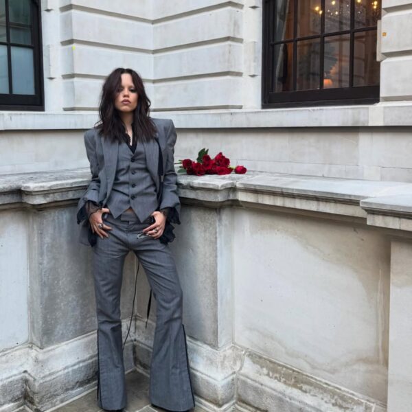 Młoda kobieta o długich, ciemnych, lekko potarganych włosach stoi oparta o kamienną balustradę w eleganckim, szarym garniturze o lekko poszerzanych nogawkach i warstwowej marynarce, z dłonią wsuniętą w kieszeń; ma wyrazisty, poważny wyraz twarzy, a obok niej na balustradzie leży bukiet czerwonych róż, tworząc kontrast z jasną, klasycystyczną elewacją budynku w tle.