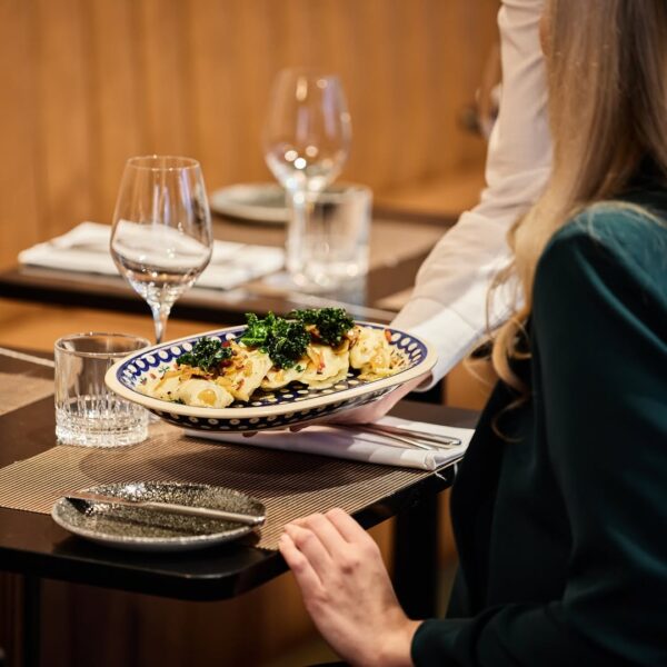Kelner podaje elegancko przygotowane danie przy stoliku, tworząc atmosferę spokojnej i uroczystej Wigilii spędzanej w restauracji. Taki moment kojarzy się z restauracją w Warszawie otwartą 24 grudnia, gdzie goście mogą celebrować świąteczną kolację w komfortowych, stylowych wnętrzach.