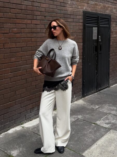Stylizacja z miękkim szarym swetrem, jasnymi spodniami i skórzaną torbą pokazuje, jak luksusowe trendy w 2026 roku łączą komfort z wyrafinowanym detalem. Kontrast faktur, subtelna koronka i stonowana paleta kolorów podkreślają odejście od surowego minimalizmu na rzecz bardziej zmysłowej elegancji.