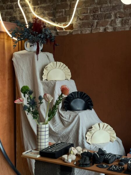 Wnętrze jednego z warszawskich concept store'ów do szukania prezentów, z ekspozycją torebek w plisowanej formie i minimalistyczną biżuterią. Na pierwszym planie stoi wazon z kwiatami i drobne akcesoria ułożone na stole. Całość utrzymana jest w spokojnej, estetycznej aranżacji.