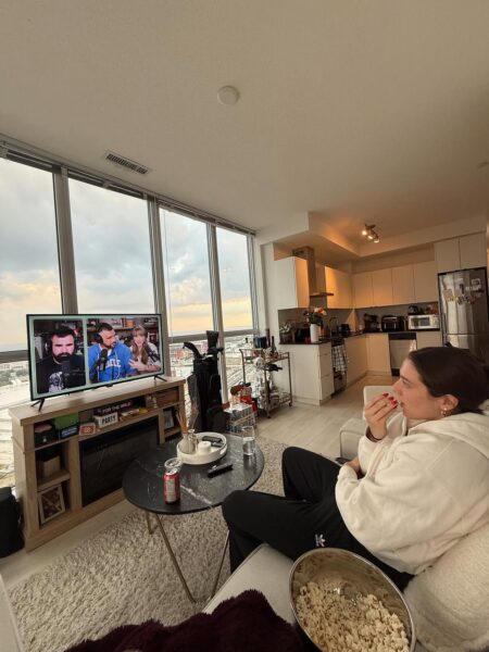 Kobieta siedzi na kanapie w nowoczesnym mieszkaniu i ogląda program na telewizorze, trzymając miskę z popcornem. Wnętrze jest jasne, z dużymi oknami i widokiem na miasto, co podkreśla domowy, wieczorny klimat. To kadr pokazujący spokojny czas spędzony przed ekranem, bez pośpiechu i planów na wyjście.