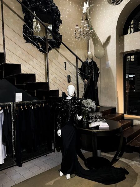 Manekin w czarnej, awangardowej stylizacji stoi w teatralnie oświetlonym wnętrzu, pełnym luster, rzeźb i dekoracyjnych detali. To przykład estetyki, którą można spotkać w concept store'ach w Warszawie, zwłaszcza tych stawiających na mocne modowe akcenty. Kompozycja podkreśla artystyczny charakter przestrzeni.