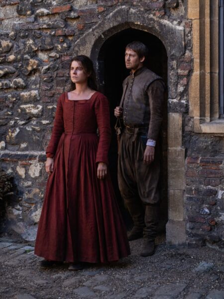 Kadry z planu filmowego, ukazujący dwoje aktorów w kostiumach z epoki renesansu lub wczesnej nowożytności, stojących przed kamiennym, łukowatym przejściem. Kobieta w pierwszym planie nosi długą, elegancką suknię w kolorze ciemnej czerwieni, a jej poważne spojrzenie oraz kostium mężczyzny w brązowych, skórzanych elementach, sugerują, że może to być jeden z historycznych dramatów.