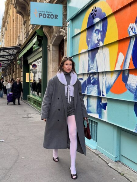 Modelka spaceruje miejskim chodnikiem, ubrana w długi, szary płaszcz z futrzanym kołnierzem w kolorze jasnego fioletu lub szarości, co stanowi mocny akcent, który może wskazywać na mikrotrendy na zimę 2026 w zakresie kolorowych akcentów wierzchnich okryć. Pod płaszczem widać krótki top i szorty w odcieniu szarości lub w kratę, kontrastujące z wyrazistymi, jasnoróżowymi rajstopami i eleganckimi, lakierowanymi butami na niewysokim obcasie. Całość prezentuje odważne połączenie tekstur i kolorów na tle eklektycznej fasady budynku z intensywnym, graficznym muralem.