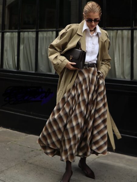 Kobieta w klasycznej stylizacji utrzymanej w brązach i beżach, z długim, rozkloszowanym trenczem w kolorze khaki i masywną spódnicą w dużą kratę, prezentuje idealny przykład elegancji, która kształtuje mikrotrendy na zimę 2026 w obszarze maxi fasonów. Zestaw dopełnia biała koszula, ciemna torebka o sztywnym fasonie oraz szpiczaste czółenka, tworząc ponadczasowy, lecz świeży look.