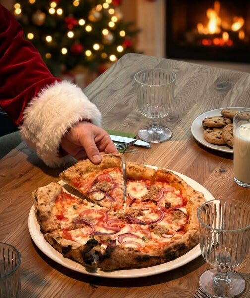 Pizza podawana na drewnianym stole w świątecznym otoczeniu, z choinką i kominkiem w tle, buduje ciepły, nieformalny klimat wspólnego posiłku. Taki kadr pasuje do restauracji otwartej w Boże Narodzenie 2025 w Warszawie, gdzie 25 grudnia można spędzić pierwszy dzień świąt przy swobodnym, rodzinnym jedzeniu.