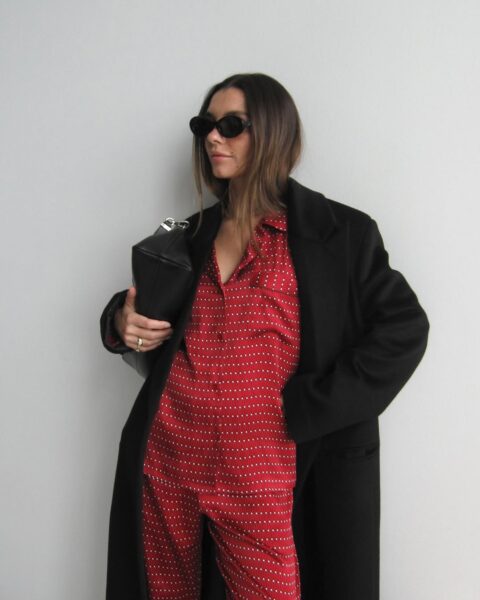 Kobieta w stylowym, miejskim looku. Czerwony, wzorzysty komplet (prawdopodobnie jedwabna piżama) wystylizowany jako dzienny strój, narzucony długim, czarnym płaszczem. Akcesoria to czarne okulary przeciwsłoneczne i elegancka torebka typu kopertówka. Zdjęcie ilustruje, jak luksusowe piżamy na 2026 rok z naturalnych tkanin wykraczają poza sypialnię, stając się elementem mody ulicznej.