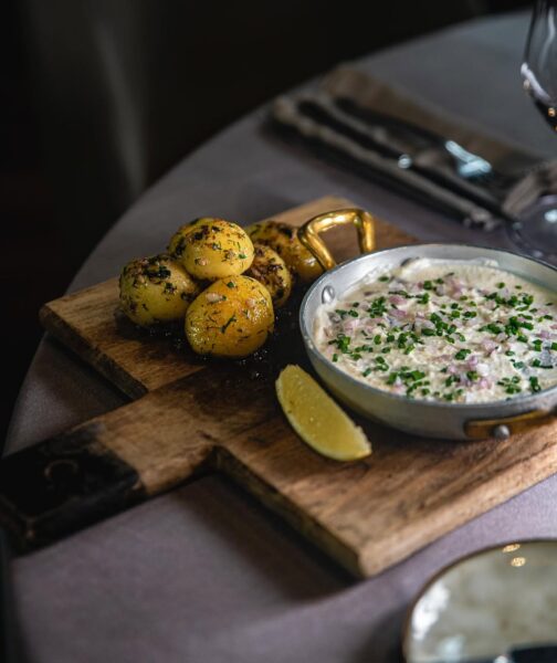 Danie podane na drewnianej desce, z ziemniakami i kremowym sosem, przywołuje skojarzenia z tradycyjną Wigilią w nowoczesnym wydaniu serwowaną w restauracji. Taki kadr pasuje do restauracji w Warszawie otwartej 24 grudnia, gdzie świąteczne smaki są podawane w eleganckiej, restauracyjnej formie.