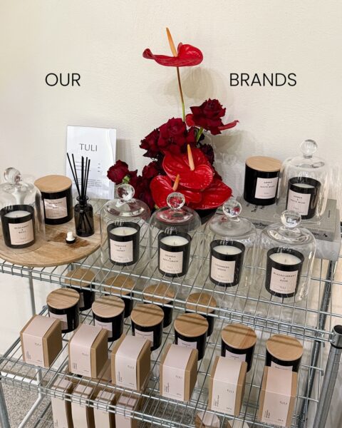 Półka z eleganckimi świecami zapachowymi Tuli, ustawionymi pod szklaną kopułą, obok bukietu czerwonych kwiatów. Tego typu starannie wyeksponowane dodatki często pojawiają się w warszawskich concept store'ach do szukania prezentów. Zdjęcie pokazuje minimalistyczną, estetyczną aranżację.