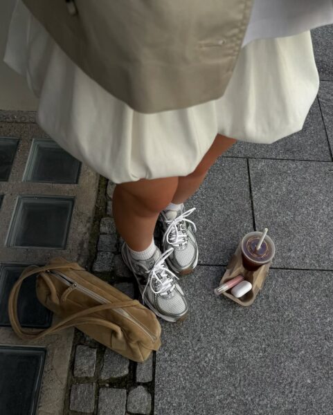 djęcie zrobione z góry przedstawia osobę stojącą na szarym chodniku w białej, obszernej spódniczce mini oraz srebrno-białych sneakersach z dużą ilością siatki i detali. Obok stóp leży beżowa zamszowa torebka i napój, co nadaje ujęciu modowy i miejski klimat. Ten look idealnie wpisuje się w modę na techniczne buty z lat 2000., która sprawiła, że właśnie te sylwetki należą obecnie do najmodniejszych modeli New Balance.