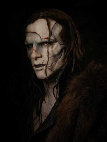 Portret osoby z imponującą charakteryzacją w stylu mrocznego fantasy lub horroru; widoczne są blade, popękane rysy twarzy oraz ciemne, długie włosy i futrzany płaszcz. Ten przerażający wygląd i mistrzostwo makijażu budzą ciekawość, czy ten typ estetyki zdominuje również nadchodzące najlepsze filmy 2025 z gatunku grozy i widowiska. Mrok i intensywność tego kadru doskonale oddają kinową atmosferę, którą fani uwielbiają i której oczekują po wielkich premierach.