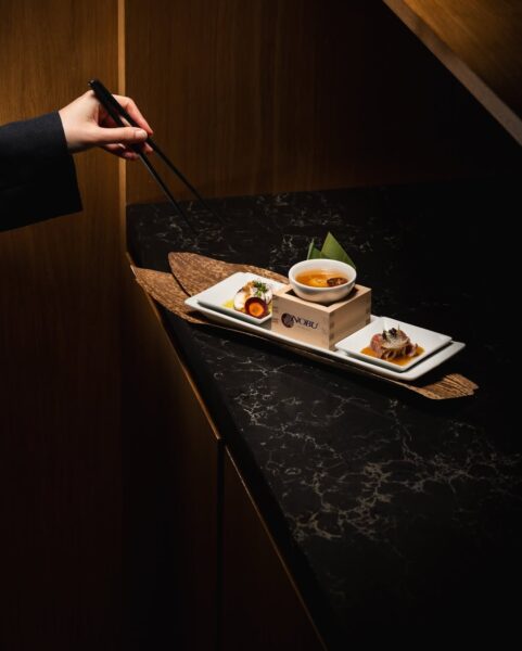 Minimalistycznie podane dania na eleganckim blacie i dłonie sięgające po potrawę pałeczkami budują atmosferę wyjątkowej kolacji w restauracji podczas Wigilii. Taki kadr pasuje do restauracji w Warszawie otwartej 24 grudnia, oferującej świąteczne menu w nowoczesnym, kameralnym wnętrzu.