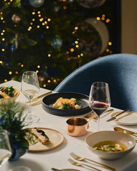 Elegancko nakryty stół w restauracji, z odświętnymi daniami, kieliszkami wina i świątecznym klimatem podkreślonym choinką w tle, przywodzi na myśl restaurację otwartą w Wigilię, gotową przyjąć gości szukających wyjątkowej kolacji. Ciepłe światło i staranna aranżacja sugerują wysokiej klasy restaurację w Warszawie, idealną na spokojne, uroczyste świętowanie poza domem w 2025 roku.