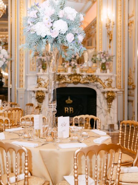Luksusowo udekorowana sala balowa z okrągłymi stołami nakrytymi złotymi obrusami, kryształowymi kieliszkami i świecznikami. Na środku stołu wysoka kompozycja kwiatowa z pastelowych róż i hortensji, a w tle kominek i bogato zdobione sztukaterie.