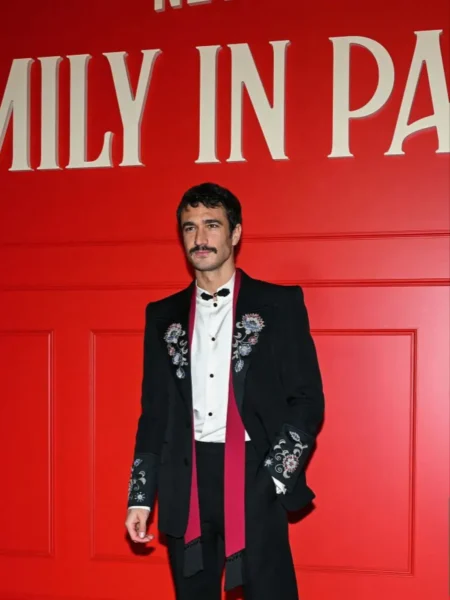 Mężczyzna w eleganckim czarnym garniturze z haftowanymi zdobieniami i białej koszuli pozuje na tle czerwonej ściany z napisem „Emily in Paris” podczas wydarzenia premierowego w Paryżu; stylizacja łączy klasyczną elegancję z modowym detalem haute couture.