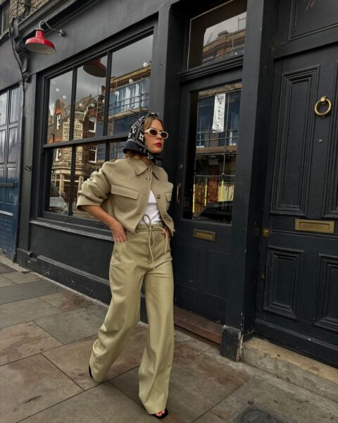 Zdjęcie przedstawia osobę idącą miejskim chodnikiem w stylizacji opartej na odcieniach khaki: krótkiej kurtce o pudełkowym kroju oraz szerokich, lejących spodniach z wiązaniem w pasie. Całość podkreślają retro okulary i apaszka z graficznym wzorem, dodające lookowi charakteru. Ujęcie idealnie wpisuje się w trend khaki coded z Pinterest Predicts 2026, który promuje neutralne palety w odważnych, nowoczesnych formach.