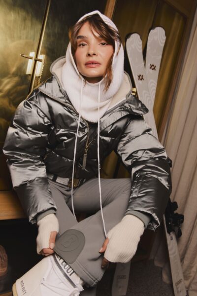 Kobieta w srebrnej, pikowanej kurtce puchowej z wysokim połyskiem, białym golfem i dopasowanymi spodniami, siedząca we wnętrzu górskiego chalet; stylizacja zimowa inspirowana modą après-ski, w tle narty i drewniane wykończenia.