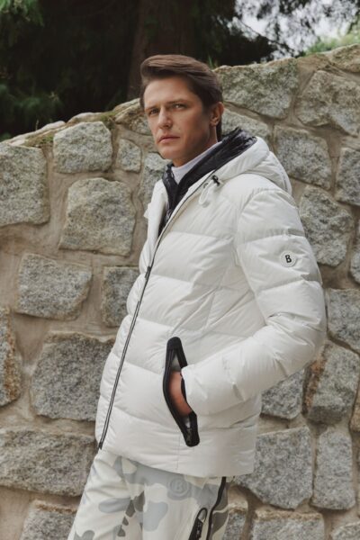 Mężczyzna w białej kurtce puchowej o sportowym kroju, stojący przy kamiennym murze w plenerze; stylizacja zimowa z elementami outdoor, neutralna paleta kolorów i minimalistyczny design.