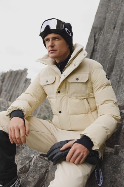 Mężczyzna w jasnym, kremowym kombinezonie narciarskim z pikowaną kurtką i czarnymi akcesoriami, siedzący na skałach w górskim krajobrazie; funkcjonalna moda zimowa inspirowana sportami alpejskimi.