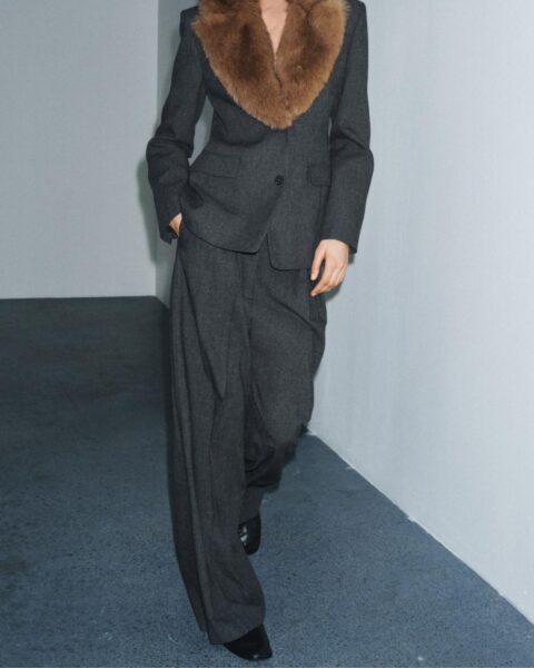 Modelka w długiej, oliwkowozielonej sukience maksi z aksamitu, z głębokim dekoltem w kształcie litery V i cienkimi ramiączkami, idealnej na wieczorowe wyjście. Kobieta z platynowym, krótkim uczesaniem i wyrazistym makijażem, stojąca na białym tle, ma skrzyżowane ramiona i długie, wiszące kolczyki.