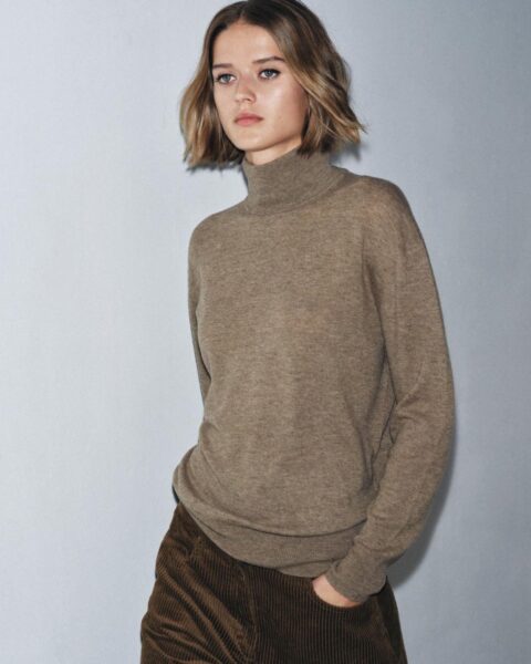 Kobieta w eleganckim, szarym garniturze z szerokimi nogawkami i taliowaną marynarką. Stylizacja uzupełniona jest dużym, brązowym, sztucznym futrem na kołnierzu. Modelka idzie korytarzem z ręką w kieszeni spodni.