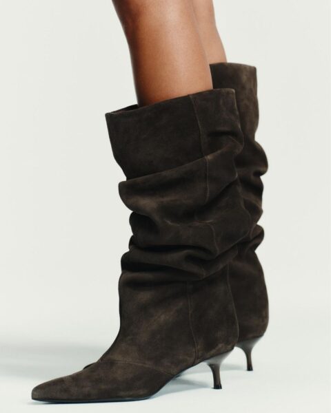 Kobieta w ciepłej, jesienno-zimowej stylizacji: beżowo-brązowy, luźny golf z wełny lub kaszmiru oraz szerokie, brązowe spodnie sztruksowe z wysokim stanem. Modelka z krótkimi, falowanymi włosami, z ręką w kieszeni.