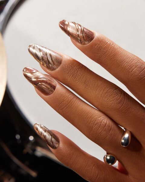 Zbliżenie na dłoń z długimi paznokciami, pomalowanymi metalicznym, miedziano-złotym lakierem hybrydowym z brokatem i falistymi, reliefowymi zdobieniami w jaśniejszym, złotym odcieniu. Na palcu serdecznym widoczny jest srebrny pierścionek z dwoma kulkami. W tle widoczna jest metalowa powierzchnia, prawdopodobnie instrumentu.