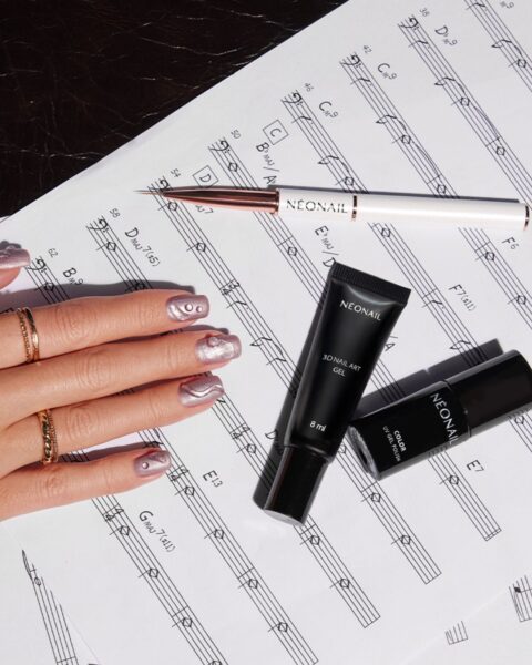 Ujęcie z góry na dłoń z krótkimi paznokciami, pomalowanymi metalicznym, brokatowym lakierem hybrydowym w odcieniu bladego różu lub liliowego beżu. Na paznokciach widoczne są subtelne, faliste wzory 3D w jaśniejszym, błyszczącym kolorze. Na palcu serdecznym i małym noszone są złote pierścionki. Dłoń spoczywa na nutach, obok leżą czarna tubka NEONAIL 3D NAIL ART GEL, czarna buteleczka NEONAIL COLOR UV GEL POLISH oraz pędzelek do zdobień z białą rączką.