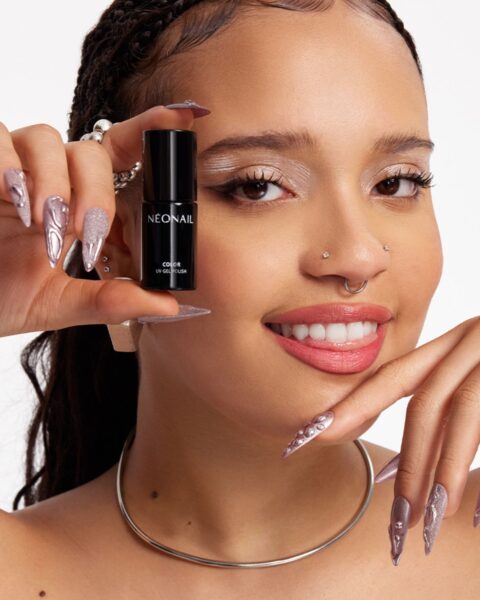 Zbliżenie na uśmiechniętą, młodą kobietę z długimi, plecionymi włosami, która trzyma czarną buteleczkę lakieru hybrydowego NEONAIL COLOR UV GEL POLISH tuż przed swoją twarzą. Ma ona intensywny makijaż oczu w odcieniach srebra i bieli oraz pomalowane usta błyszczącym różem. Na dłoni widoczny jest ekstremalny, spiczasty manicure w kolorze błyszczącej lawendy, ze srebrnymi i brokatowymi akcentami oraz kryształkami. Kobieta ma na sobie srebrną kolie i kolczyk w nosie.