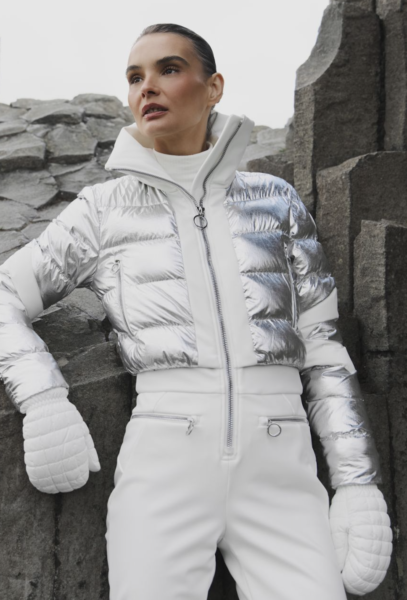 Kobieta w srebrno-białym kombinezonie zimowym z połyskującą, krótką kurtką puchową i wysokim kołnierzem, stojąca w skalnym otoczeniu; nowoczesna, luksusowa moda narciarska w jasnej tonacji.
