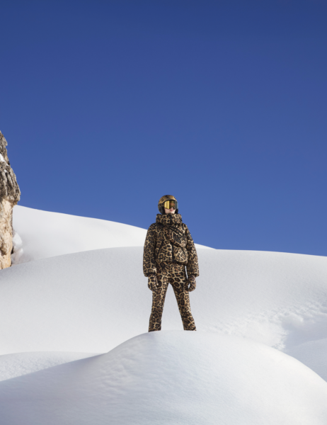 Szeroki kadr: sylwetka w komplecie narciarskim w panterkowy wzór, stojąca na zaśnieżonym wzniesieniu; kontrast pomiędzy fakturą śniegu, intensywnym printem i czystym, niebieskim niebem.