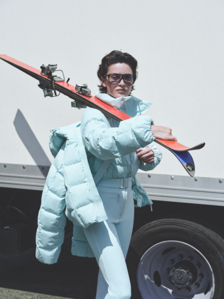 Modelka w pastelowym, błękitnym komplecie narciarskim z puchową kurtką i dopasowanymi spodniami, niosąca narty na ramieniu; stylizacja utrzymana w estetyce retro sport na tle technicznej infrastruktury.