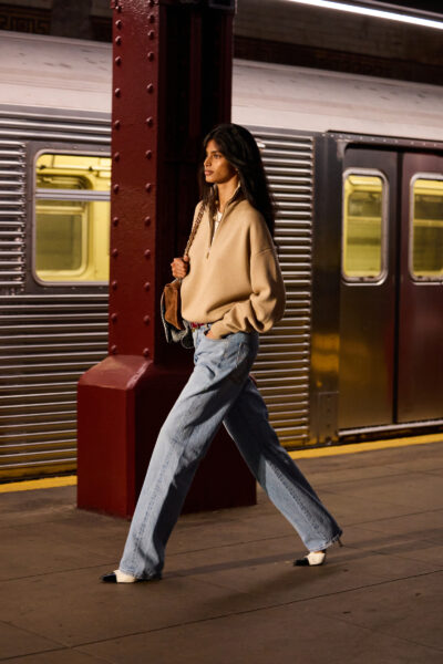 Modelka w casualowo-eleganckiej stylizacji Chanel — beżowej bluzie typu półgolf i szerokich jeansach, z torebką na złotym łańcuchu przewieszoną przez ramię; dynamiczny krok podkreśla sylwetkę przemieszczającą się wzdłuż wagonu metra.