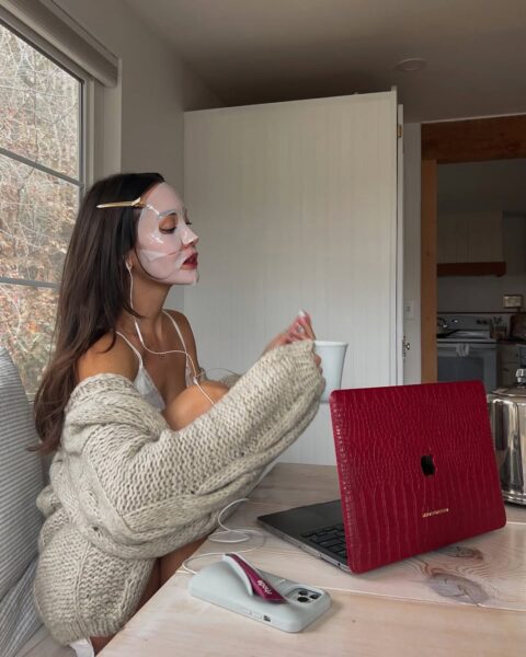 Filmy na karnawał: Młoda kobieta w swetrze siedzi przy stole obok czerwonego laptopa i pije kawę, korzystając jednocześnie z maski w płacie na twarzy. Relaksuje się w jasnym wnętrzu przy dużym oknie, co nadaje scenie spokojny, poranny charakter. Jest to idealny przykład, jak maski kolagenowe można wpleść w codzienny harmonogram pracy lub odpoczynku.