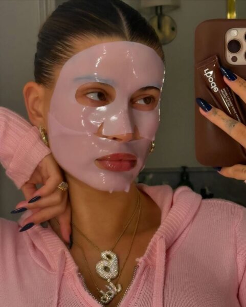 Modelka Hailey Bieber w różowej bluzie z kapturem robi selfie w lustrze, trzymając w dłoni telefon. Na twarzy ma gładką, różową maskę w płacie, która intensywnie nawilża skórę. Wielu uważa te najlepsze maski kolagenowe za sekret jej promiennego wyglądu i idealnie gładkiej cery.