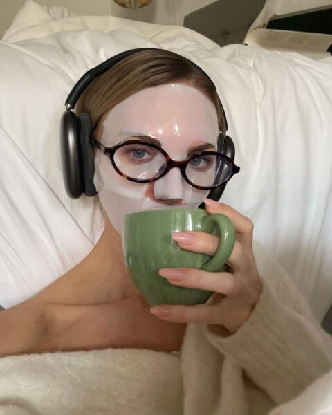 Kobieta leży na białej pościeli, relaksując się z kubkiem zielonej herbaty i słuchawkami na uszach. Na jej twarzy widać transparentną, nawilżającą maskę w płacie, która zapewnia głębokie odżywienie. Wykorzystanie produktu z kategorii maski kolagenowe to świetny sposób na domowy relaks i regenerację skóry.