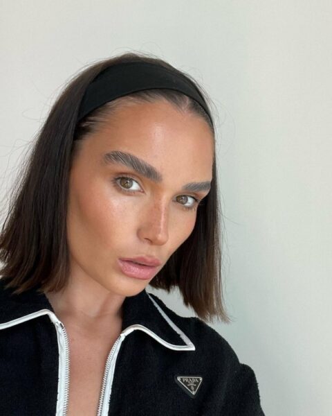 Zdjęcie portretowe przedstawia kobietę z eleganckim makijażem, ubraną w czarną kurtkę z białym kołnierzem, a na jej głowie widoczna jest szeroka, czarna opaska. Opaska podtrzymuje klasycznego, równego boba, który jest gładko zaczesany do tyłu, co podkreśla modny minimalizm i będzie jednym z wiodących trendów w kategorii modne fryzury 2026. Ten ponadczasowy, schludny styl z akcentem w postaci opaski to esencja wyrafinowanej prostoty.