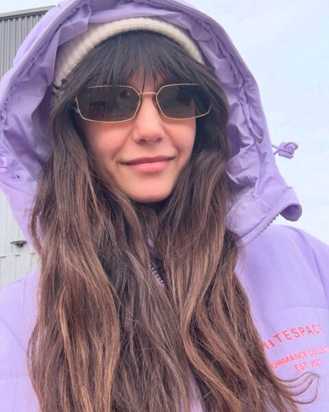 Portretowe selfie przedstawia aktorkę Ninę Dobrev w ciepłej, fioletowej kurtce narciarskiej z kapturem i w modnych okularach przeciwsłonecznych. Jej zrelaksowany uśmiech i sportowy strój gotowy na stok świetnie obrazują temat: jak gwiazdy spędzają zimę w aktywny i stylowy sposób.