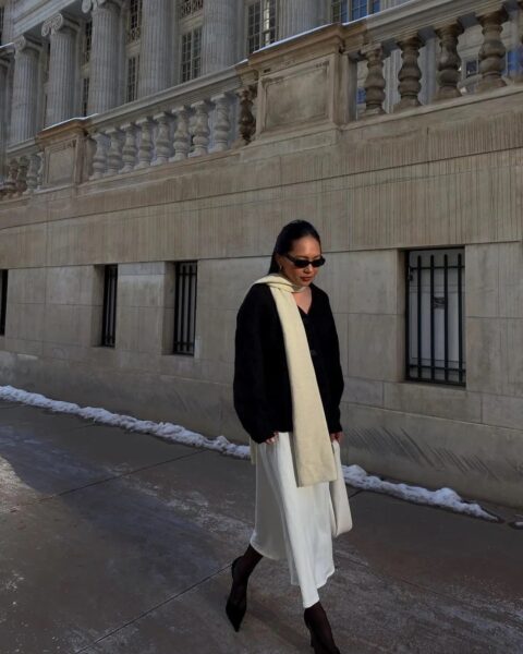 Elegancka stylizacja na chłodny dzień, prezentująca połączenie czarnego, luźnego swetra z dekoltem w serek z długą, kremową spódnicą, co idealnie wpisuje się w mikrotrendy na zimę 2026 stawiające na klasyczne kontrasty. Biały, długi szal zarzucony na ramię dodaje całości szyku, a całość dopełniają czarne szpilki i okulary przeciwsłoneczne, tworząc nowoczesny look na tle monumentalnej, kamiennej architektury.