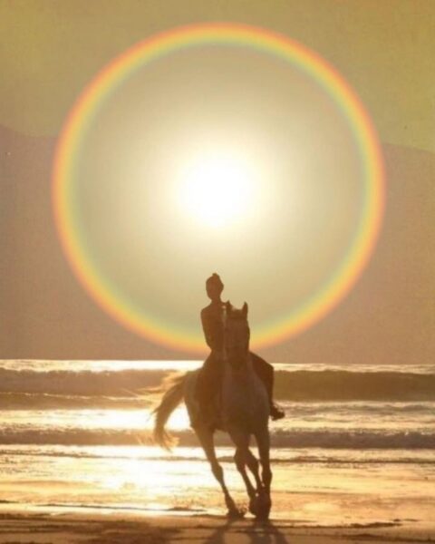 Artystyczne zdjęcie przedstawiające ciemną sylwetkę osoby jadącej na koniu brzegiem morza o zachodzie słońca, otoczoną świetlistymi, kolistymi flarami w kształcie aureoli.
