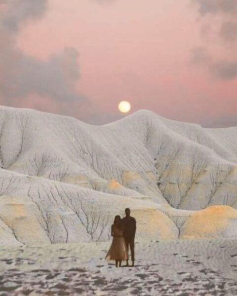 Artystyczne ujęcie pary stojącej na tle jasnych, pofalowanych wzgórz przypominających księżycowy krajobraz pod różowym niebem z pełnią księżyca – astrologia 2026.