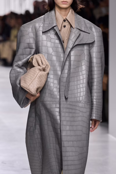Detal stylizacji z wybiegu: modelka ubrana w długi, strukturalny płaszcz w odcieniu chłodnej szarości, wykonany z tłoczonej skóry o fakturze imitującej krokodyla. Stylizację uzupełnia beżowa koszula z klasycznym kołnierzem oraz miękka, pleciona kopertówka noszona pod pachą. Ujęcie podkreśla minimalistyczną paletę barw i luksusowe wykończenie materiałów.